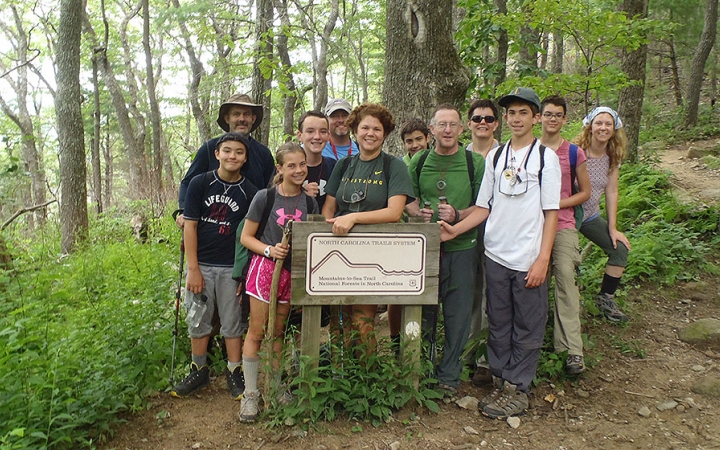 A group of parents and children pose for a photo near a sign in a green wooded area. The words on the sign are too small to read.